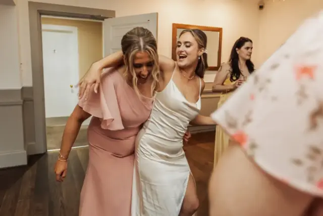 Two women, one in a pink dress and the other likely the bride in white, dance joyfully together indoors at Whirlow Brook Hall. The woman in white has her arm around the other as they smile, enjoying a wedding celebration with others in the background.