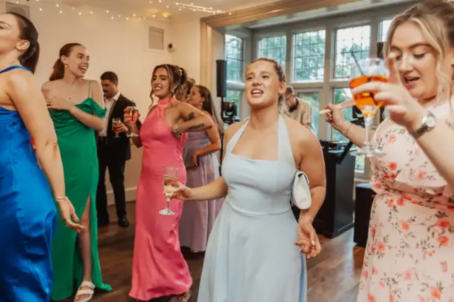 A group of women in colorful dresses dance and sing while holding drinks at a wedding celebration at Whirlow Brook Hall, with fairy lights overhead and large windows in the background.