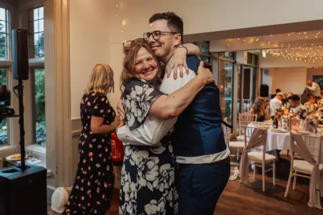 A bride in a floral dress and a man in a suit vest hug and smile at a wedding reception at Whirlow Brook Hall, with tables, lights, and other guests in the background.