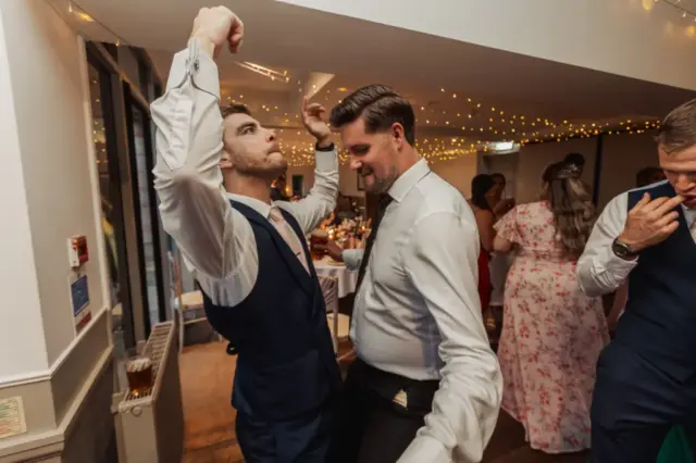 Two men in dress shirts and vests dance enthusiastically at a wedding reception, one raising his arms and the other smiling. Guests in formal attire and string lights fill the background at Whirlow Brook Hall, creating a festive atmosphere.