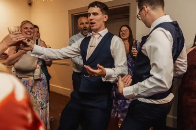 A group of people dressed in formal attire dance and laugh together at a wedding at Whirlow Brook Hall. Two men in vests are in the center, appearing animated, while others, including the bride, smile and watch in the background.