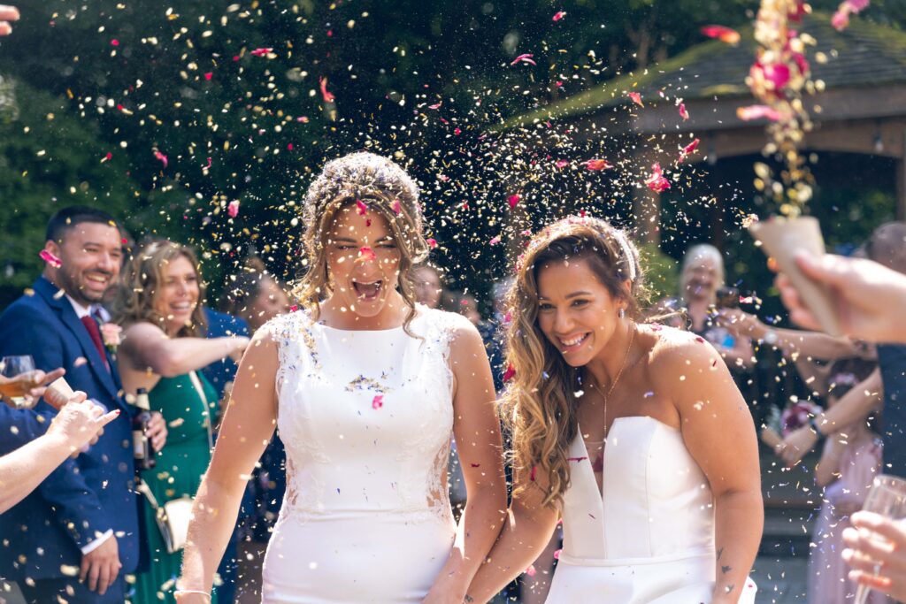 Two brides in white dresses smile and hold hands as wedding guests shower them with flower petals—some of the best wedding confetti—outdoors. People in the background cheer and celebrate in the sunlight.