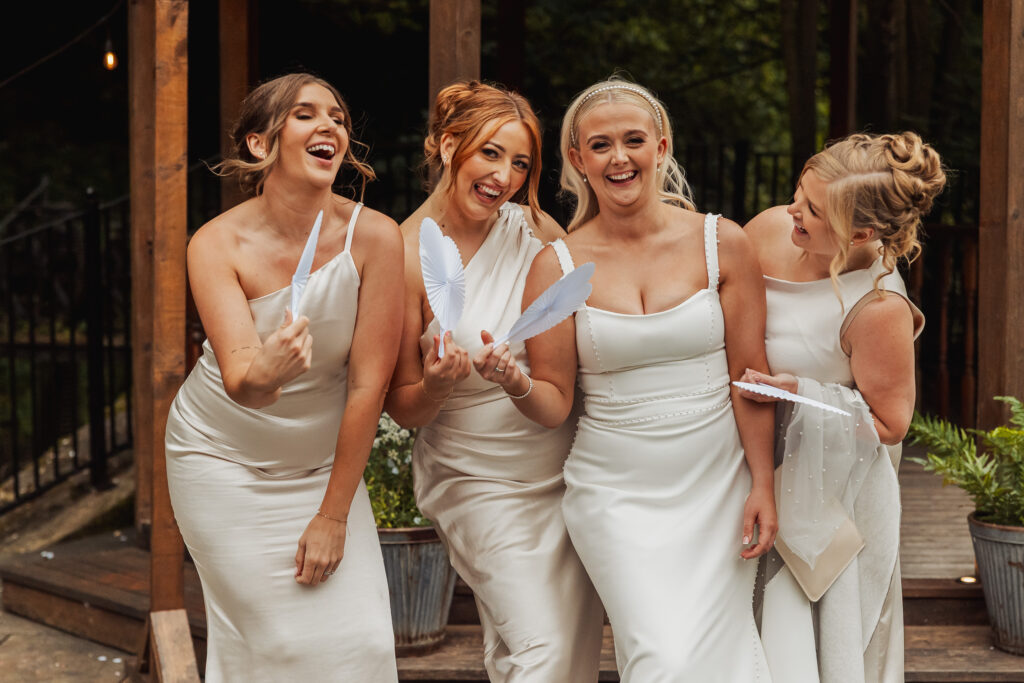 Four women in elegant light-colored dresses stand outside, smiling and laughing. Three hold white feathers, and one holds a small clutch. The scene is joyful and celebratory, possibly at a wedding or special event.
