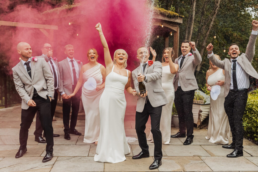 A joyful wedding party celebrates outdoors; the bride and groom lead the group, popping champagne as pink smoke from Smoke Bombs wedding effects fills the air. Bridesmaids in white dresses and groomsmen in grey jackets cheer enthusiastically.