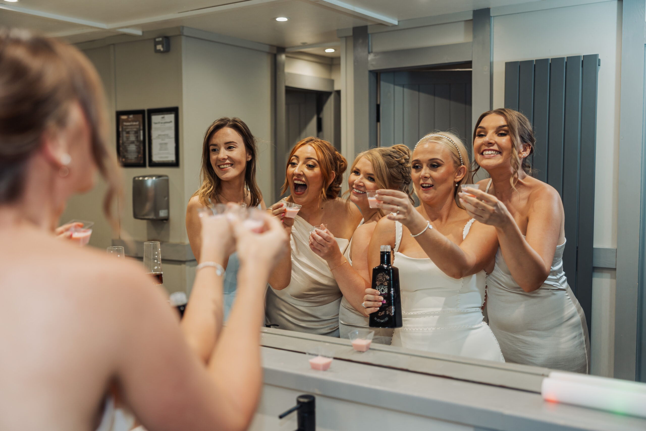Five women in light dresses smile and raise shot glasses in a toast, celebrating together in front of a mirror—perfect bridesmaid photo inspiration. One holds a bottle, and their joyful reflections add to the brightly lit scene.