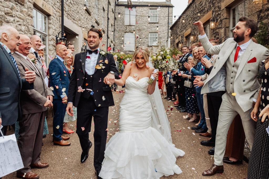 A bride and groom, smiling and holding hands, walk down an outdoor aisle as guests on both sides throw confetti—one of the best wedding traditions—between stone buildings. Everyone is dressed in formal attire and cheers fill the air.