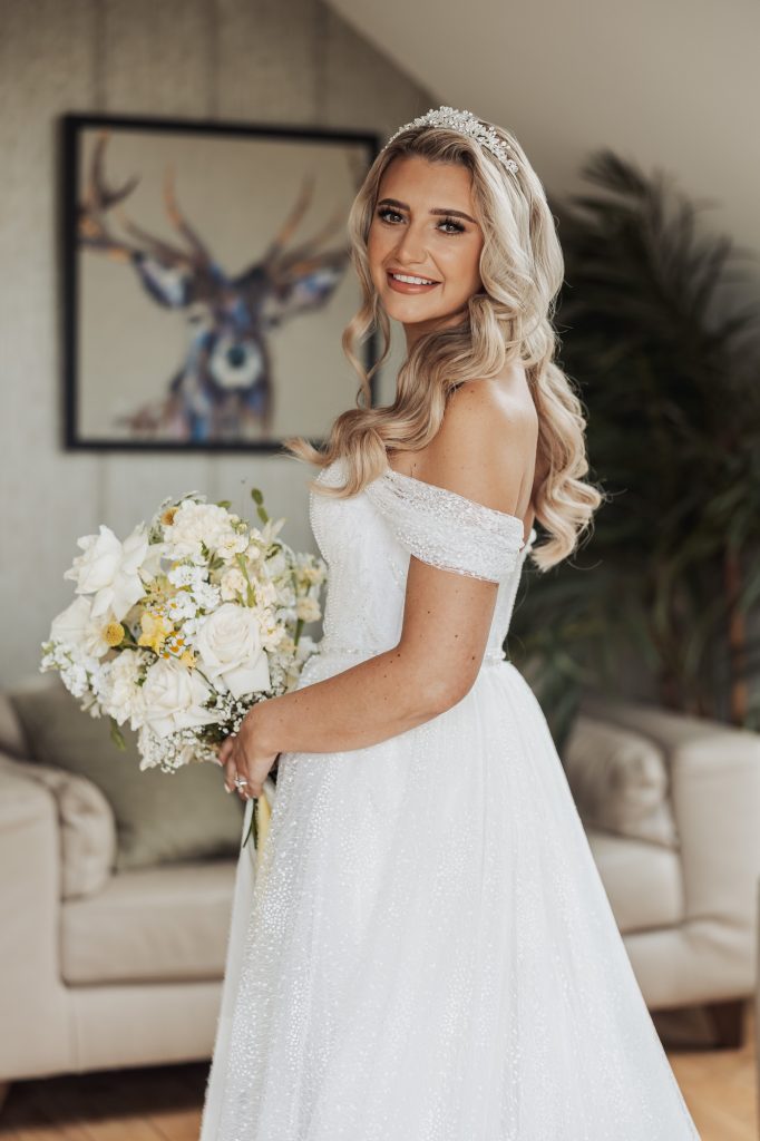 A bride with long, wavy blonde hair and a tiara smiles while holding a bouquet of white flowers, wearing an off-the-shoulder white wedding dress at her Bunny Hills wedding. A deer painting and a plant decorate the background.