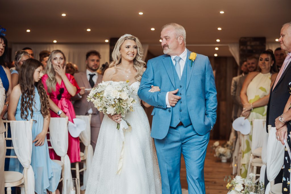 A bride in a white gown and a man in a blue suit walk arm-in-arm down the aisle, smiling, as guests watch their Bunny Hills wedding ceremony indoors.
