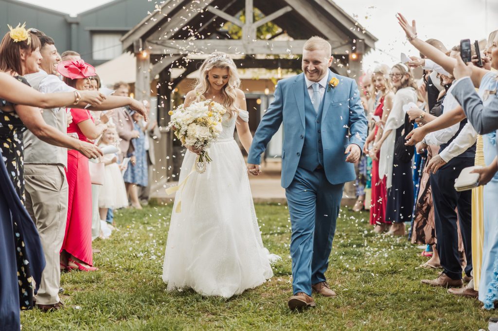 A bride and groom walk hand in hand outdoors at their Bunny Hills wedding, smiling as guests throw confetti. The crowd lines both sides, celebrating in front of a rustic wooden structure on a sunny day.