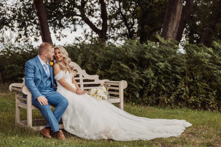 At their Bunny Hills wedding, a bride in a white gown and groom in a blue suit sit on a wooden bench outdoors, surrounded by greenery. The bride smiles with her bouquet, her dress elegantly trailing onto the grass.