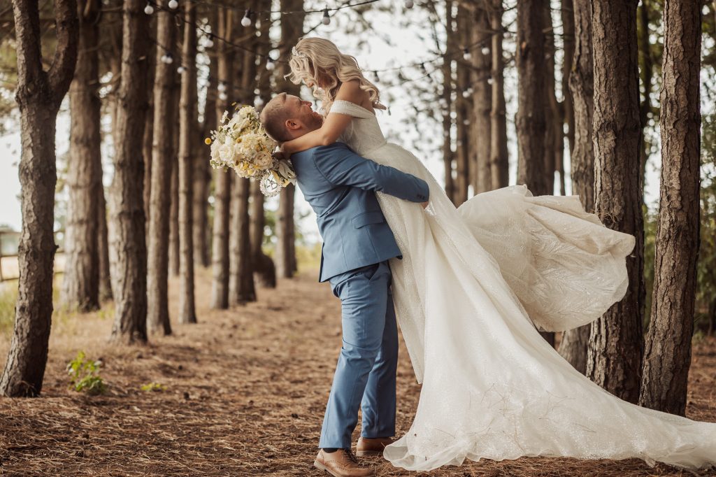 A groom in a blue suit lifts his bride in a white wedding dress and veil among tall pines at their Bunny Hills wedding. The bride holds a bouquet as they smile at each other, surrounded by the enchanting forest setting.