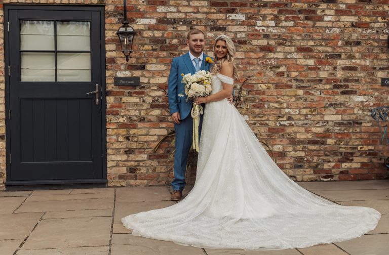A bride in a white gown with a long train stands beside her groom in a blue suit, both smiling and holding a bouquet, celebrating their Bunny Hills wedding in front of a brick wall and a dark door.