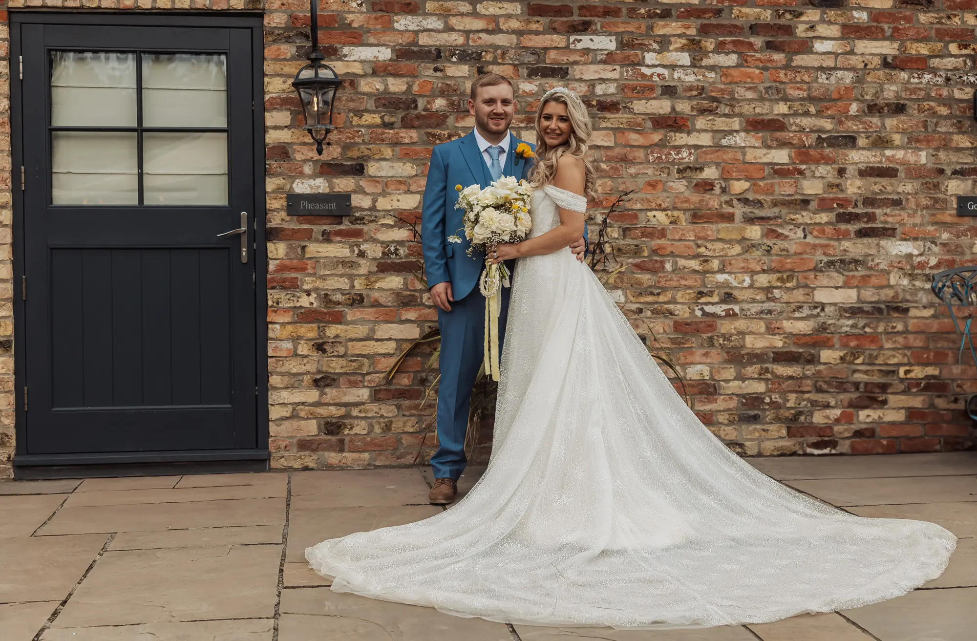 A bride in a white gown with a long train stands beside her groom in a blue suit, both smiling and holding a bouquet, celebrating their Bunny Hills wedding in front of a brick wall and a dark door.