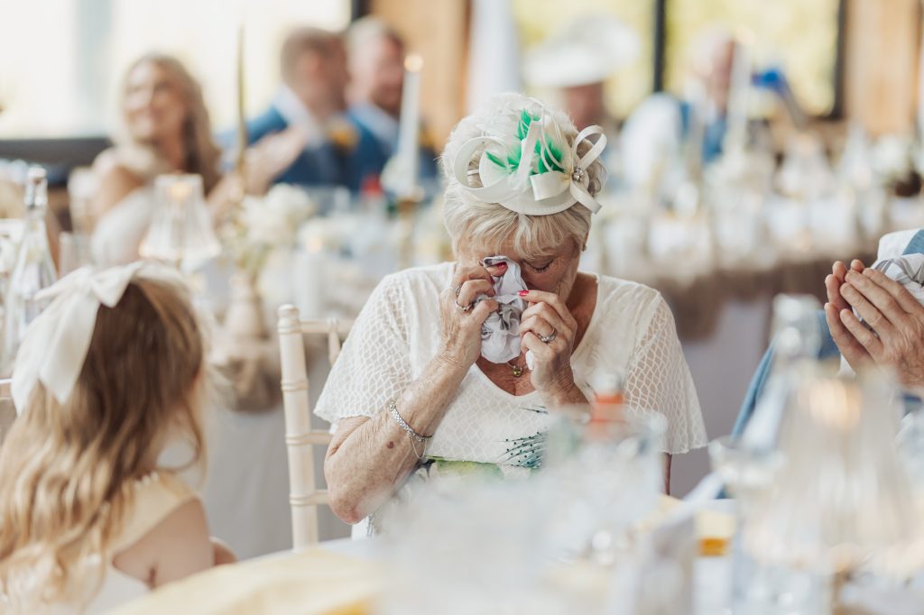 An elderly woman in a white outfit and hat wipes her eyes with a tissue at a bunny hills wedding, possibly overcome with emotion. A young girl sits nearby, while other guests are blurred in the background.