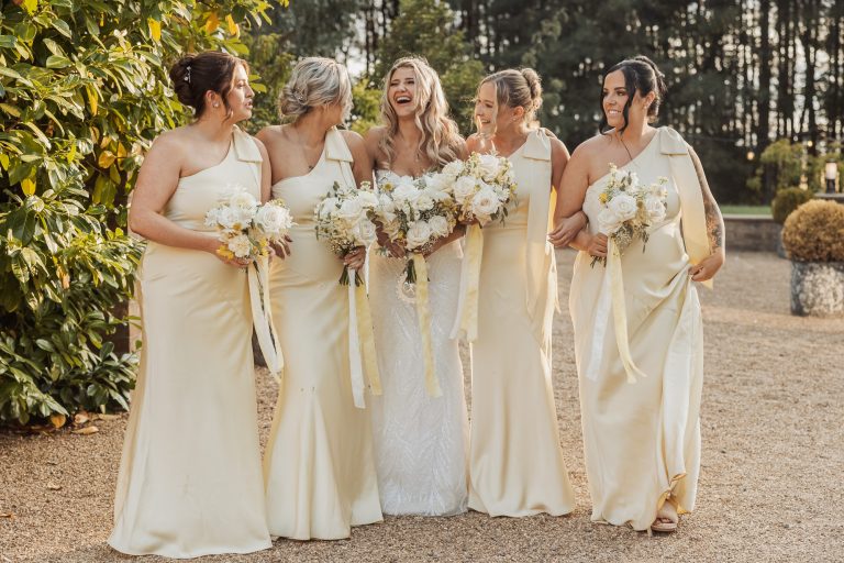 Five women stand outdoors in elegant light-colored gowns. Four bridesmaids, holding white bouquets, smile at the bride in a white gown with a larger bouquet. Lush trees and greenery suggest a charming bunny hills wedding setting.