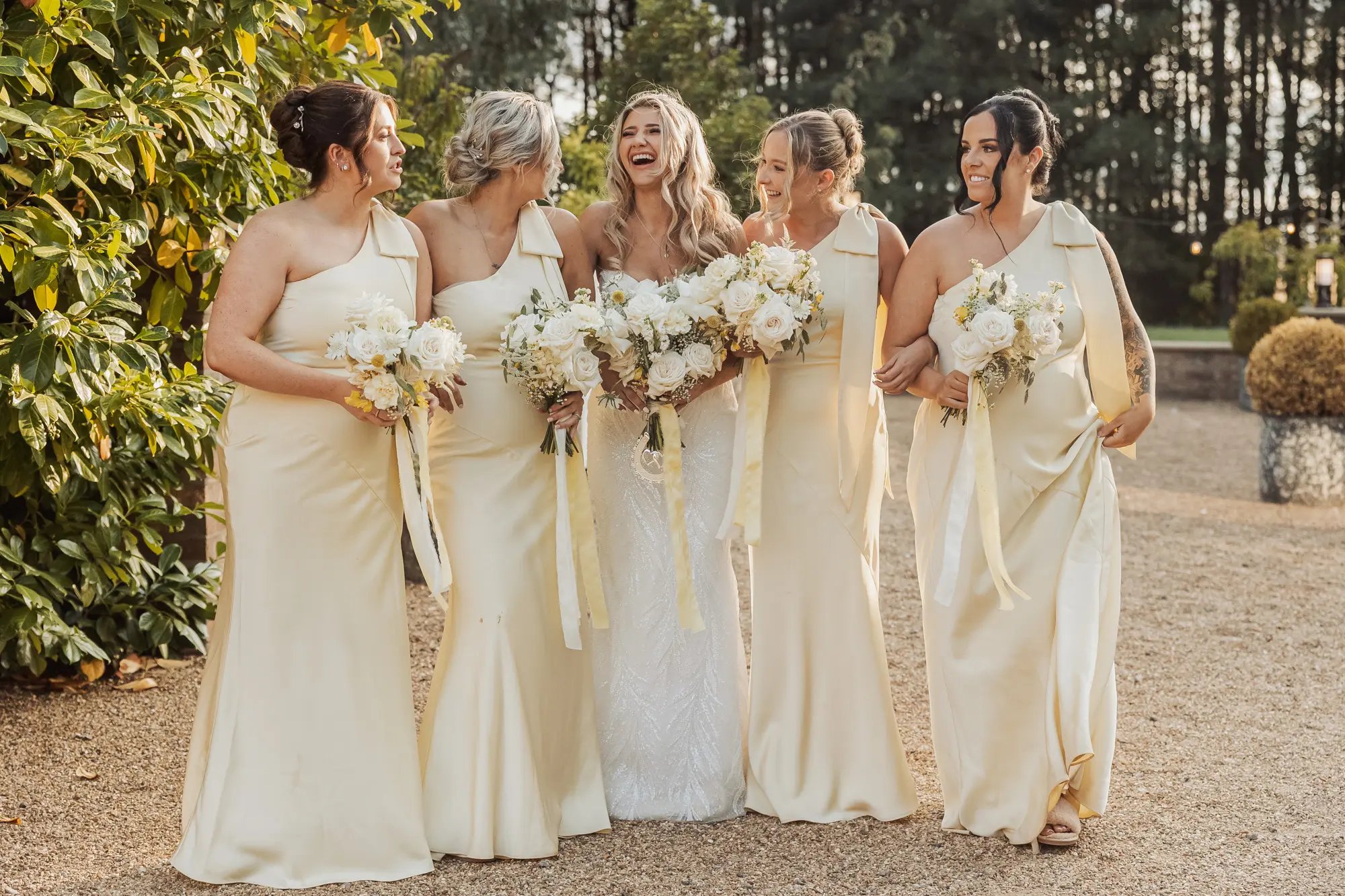 Five women stand outdoors in elegant light-colored gowns. Four bridesmaids, holding white bouquets, smile at the bride in a white gown with a larger bouquet. Lush trees and greenery suggest a charming bunny hills wedding setting.