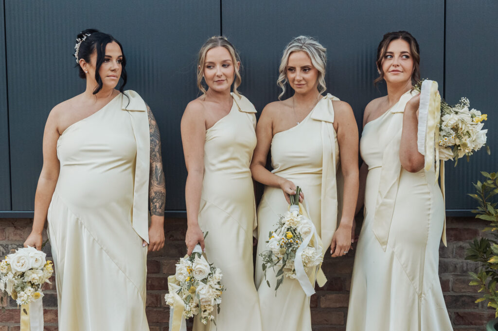Four women in matching cream, one-shoulder dresses, holding bouquets of white and yellow flowers, stand side by side against a dark wall, posing for a photo.