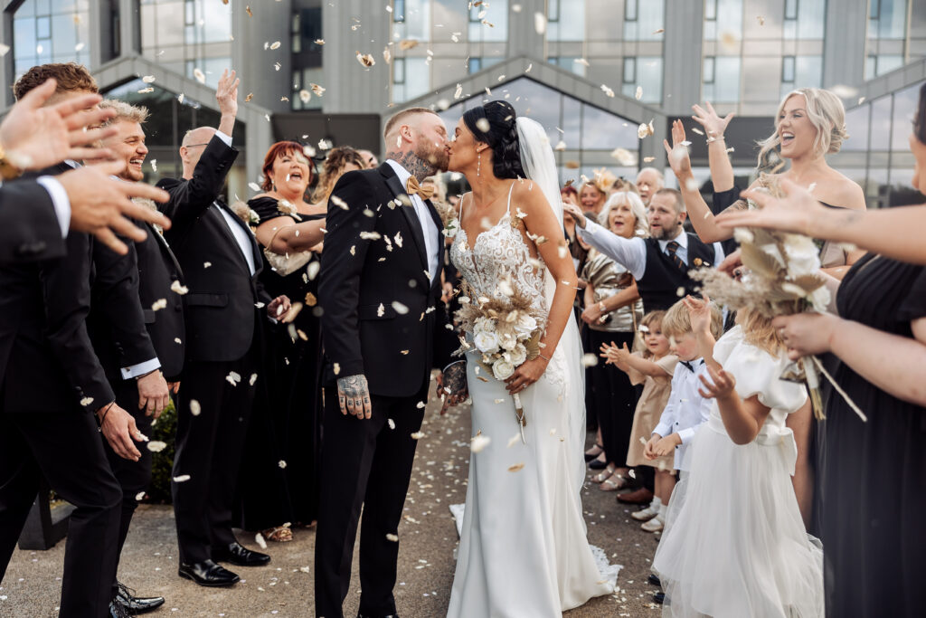 A bride and groom kiss while surrounded by guests throwing flower petals—the best wedding confetti. The couple stands at the center, smiling, with guests cheering and children holding bouquets nearby. Everyone is dressed formally.
