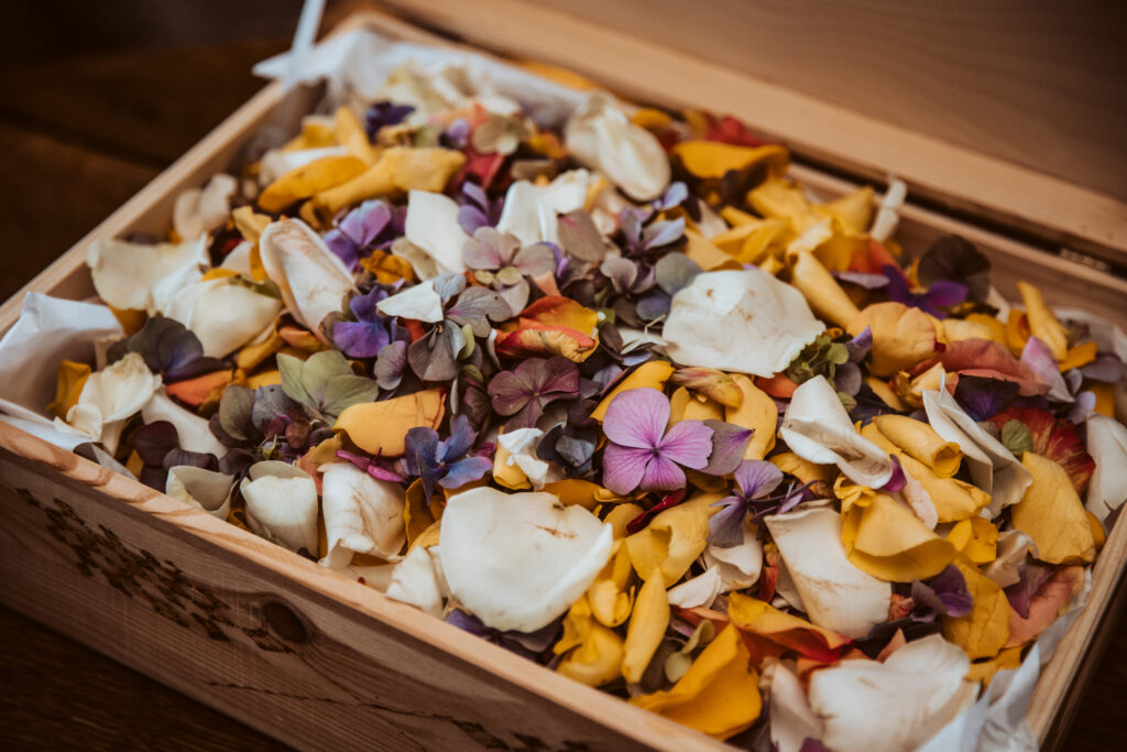 A wooden box filled with a colorful mix of dried flower petals—white, yellow, pink, and purple—arranged loosely on tissue paper. The best wedding confetti for a romantic and natural celebration.