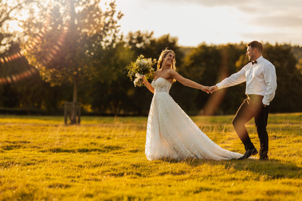 A bride in a white gown holds a bouquet and smiles while holding hands with a groom in a white shirt and black bow tie, walking together in a sunlit outdoor field at their North Yorkshire wedding.