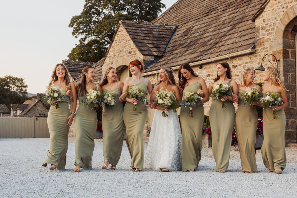 A bride in a white dress walks outdoors with eight bridesmaids in matching sage green dresses, all holding bouquets and smiling in front of the stone Tithe Barn Bolton Abbey.