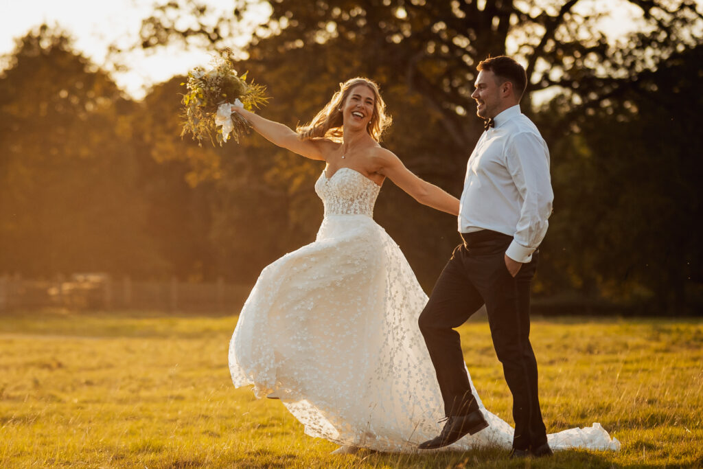 A joyful bride in a white dress twirls with her bouquet beside a smiling groom in a white shirt and bow tie, celebrating their wedding on a sunlit grassy field near the beautiful tithe barn at Bolton Abbey.