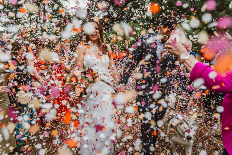 A bride and groom in wedding attire walk outdoors while guests throw colorful confetti—because knowing what confetti best suits your wedding creates a truly joyful and vibrant celebration scene.