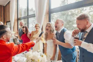 A bride and groom, along with wedding guests, laugh and sing enthusiastically into a microphone held out by a man in a red jacket at a lively indoor Bunny Hills wedding reception.