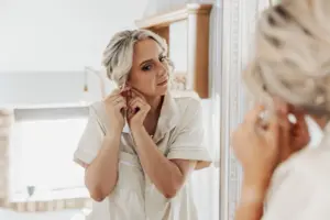 A woman with blonde hair in loose waves stands in front of a mirror, wearing cream-colored pajamas, as she puts on an earring and admires her reflection before her Bunny Hills wedding.