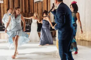 A bride in a white gown dances joyfully with guests at a Bunny Hills wedding, while a man in a blue suit sings into a microphone. The floor is lit, and everyone appears to be celebrating and having fun.