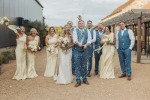 A joyful Bunny Hills wedding party poses outdoors as the groom pops a bottle of champagne, spraying bubbles, while everyone smiles and cheers. The bridesmaids wear light yellow dresses and the groomsmen sport blue suits with vests.