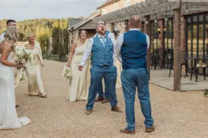 A joyful groom stands with arms wide as a man throws confetti at him outside the rustic Bunny Hills wedding venue, while the bridal party watches and laughs in the background.