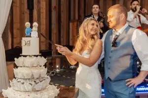A bride and groom smile at each other while cutting a tiered wedding cake topped with figurines, surrounded by cupcakes. A band performs in the background, filling this bunny hills wedding scene with warmth and festivity.