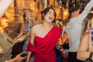 A woman in a bright red, one-shouldered dress smiles and sings while holding a drink at a lively Bunny Hills wedding, surrounded by guests celebrating joyfully around her.