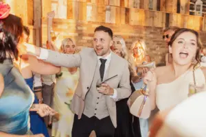 A group of people dressed in formal attire cheer and celebrate indoors at a Bunny Hills wedding. A man in a light suit holds a drink and points, while others smile, laugh, and raise their arms against a rustic wood and brick wall background.
