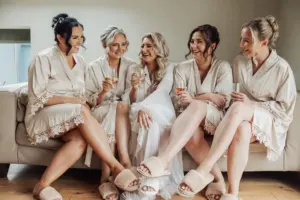 Five women in matching robes and slippers sit on a couch, smiling and holding champagne glasses, celebrating their friendship at a cozy Bunny Hills wedding.