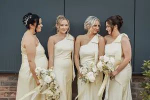Four women in matching cream-colored, one-shoulder dresses hold white floral bouquets, smiling at each other in front of a gray wall and brick background at a beautiful Bunny Hills wedding.
