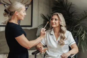 A makeup artist prepares a smiling woman with long blonde hair for her Bunny Hills wedding, as she sits in white satin pajamas, holding champagne in a cozy, plant-filled room.