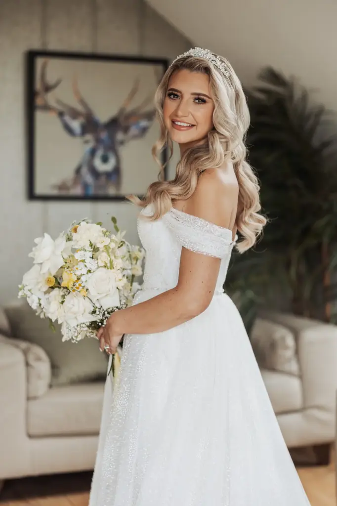 A bride with long, wavy blonde hair and a tiara smiles while holding a bouquet of white flowers, wearing an off-the-shoulder white wedding dress at her Bunny Hills wedding. A deer painting and a plant decorate the background.