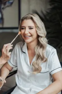 A smiling woman with long blonde hair in white satin pajamas is having makeup applied to her face with a brush, getting ready for a Bunny Hills wedding.