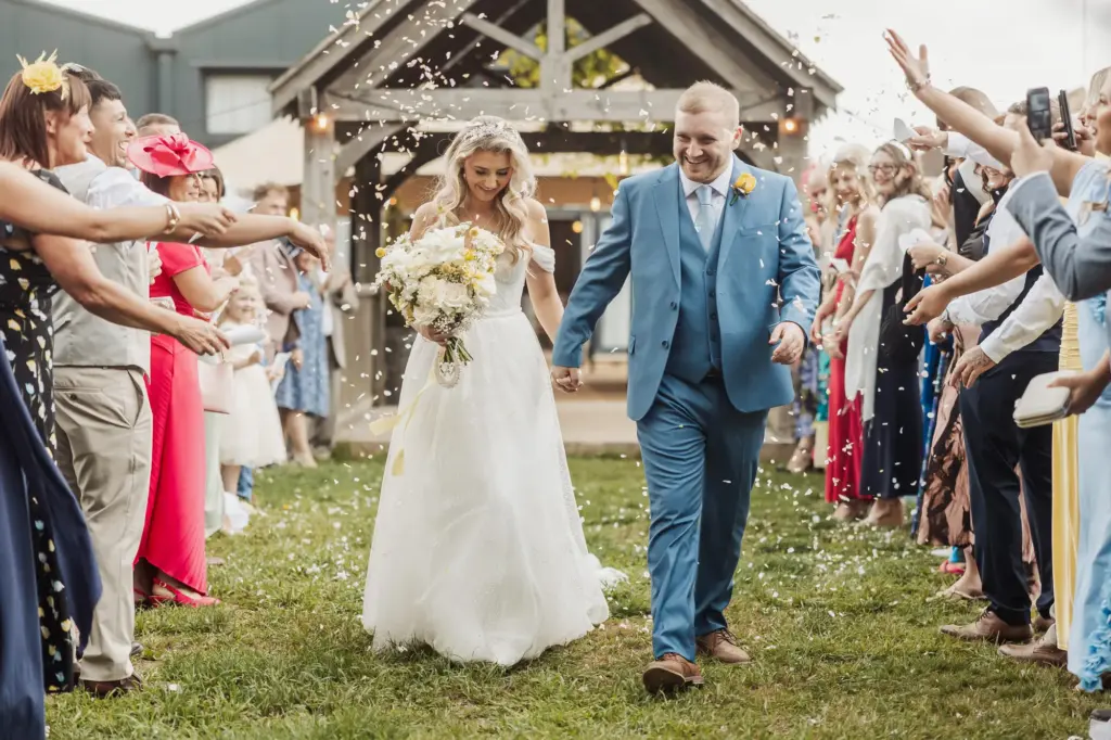 A bride and groom walk hand in hand outdoors at their Bunny Hills wedding, smiling as guests throw confetti. The crowd lines both sides, celebrating in front of a rustic wooden structure on a sunny day.