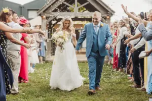 A bride and groom walk hand in hand outdoors at their Bunny Hills wedding, smiling as guests throw confetti. The crowd lines both sides, celebrating in front of a rustic wooden structure on a sunny day.