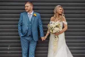 A bride in a white gown holds a bouquet and smiles at the groom, who wears a blue suit and looks away. They stand hand in hand by a dark, ribbed metal wall, capturing an elegant moment from their Bunny Hills wedding.