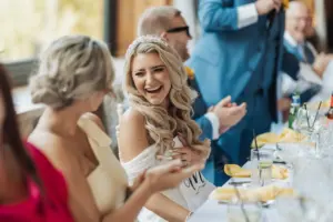 A bride in a white dress and tiara laughs joyfully while sitting at her Bunny Hills wedding reception, surrounded by elegantly dressed guests, with glasses and yellow napkins visible on the table.