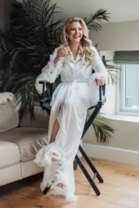 A woman in a white robe with feathered sleeves sits on a tall chair, smiling and holding a glass of champagne. She has long, wavy blonde hair and is barefoot. Green plants and a window hint at a serene Bunny Hills wedding setting.