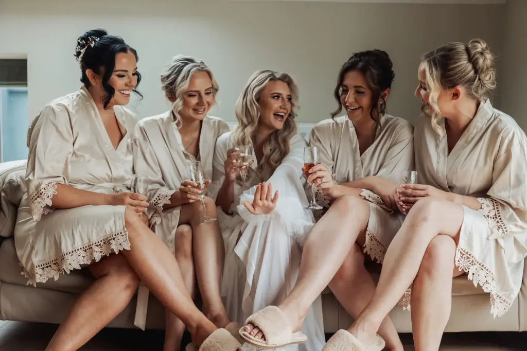 Five women in matching robes sit on a couch, smiling and laughing together while holding glasses of champagne, likely preparing for a Bunny Hills wedding or special event.