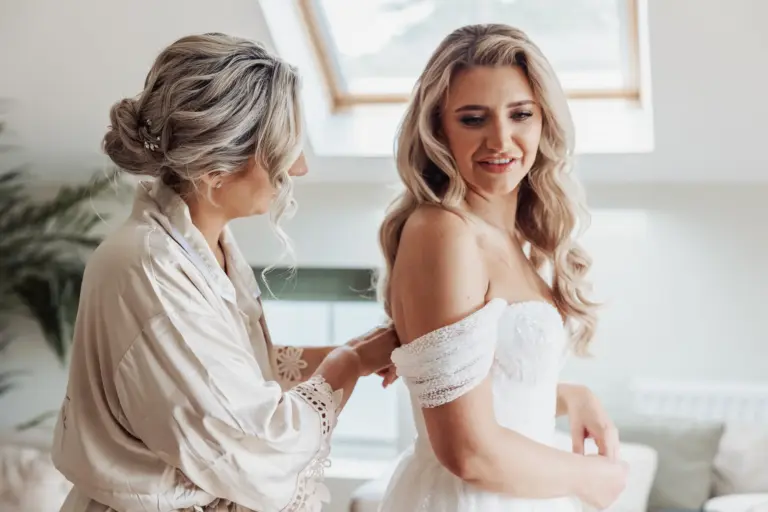 A woman in a robe helps a bride adjust her off-the-shoulder white dress. Both have long, wavy blonde hair and stand in a bright room with natural light from a skylight, preparing for a beautiful Bunny Hills wedding.