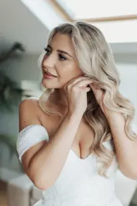A woman with long, wavy blonde hair in an off-the-shoulder white wedding dress smiles softly as she puts on an earring, standing indoors with natural light streaming in on her special Bunny Hills wedding day.