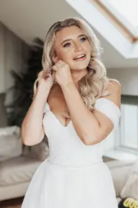 A smiling bride with long, wavy blonde hair wears an off-the-shoulder white wedding dress and puts on an earring in a bright, modern room at Bunny Hills Wedding.