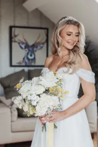A smiling bride in a white off-shoulder wedding dress holds a bouquet of white and yellow flowers at her Bunny Hills wedding. She has long, wavy blonde hair, wears a tiara, and a painting of a deer hangs on the wall behind her.